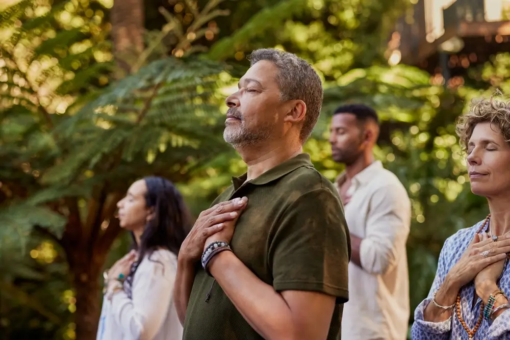 Groupe de personnes multiethniques debout à l'extérieur, les yeux fermés et les mains sur le coeur pendant une séance de méditation.