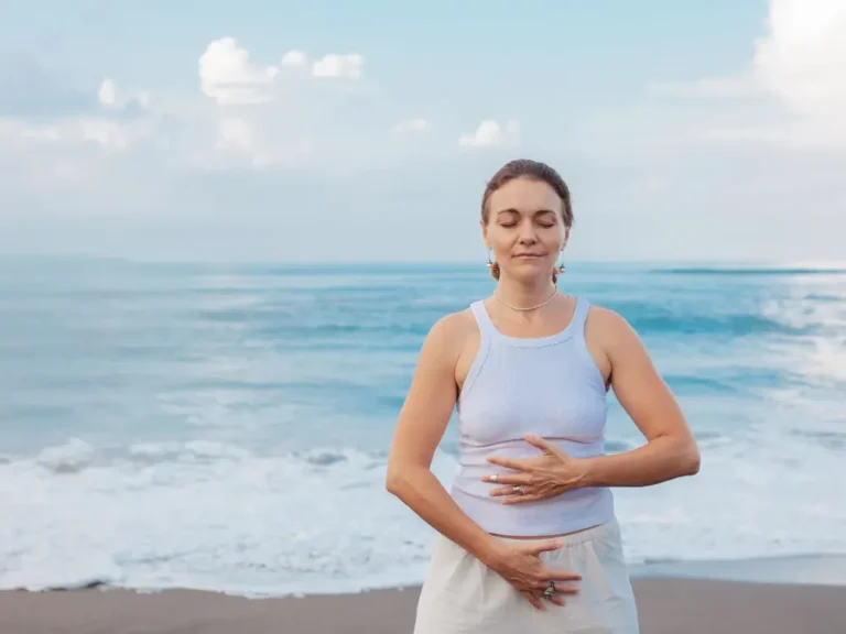 Une femme caucasienne d'âge mûr fait de l'exercice d'énergie qigong à l'extérieur sur la plage, le ciel bleu et l'océan en arrière-plan.