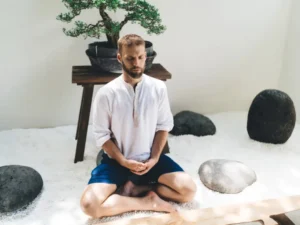 Homme assis en tailleur sur du gravier blanc à l'intérieur, méditant les yeux fermés devant un bonsaï, calme et concentré, entouré de pierres dans un espace de méditation minimaliste de style zen.