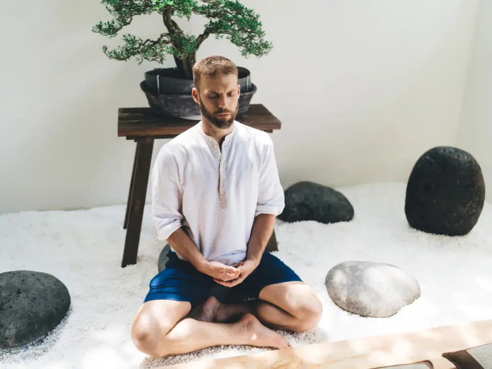 Homme assis en tailleur sur du gravier blanc à l'intérieur, méditant les yeux fermés devant un bonsaï, calme et concentré, entouré de pierres dans un espace de méditation minimaliste de style zen.