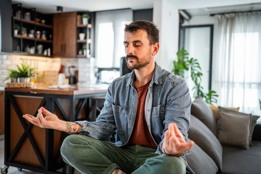 Homme pratiquant la méditation consciente dans une position détendue de lotus, assis sur un canapé dans une maison moderne, trouver la paix et l'équilibre spirituel dans sa routine quotidienne.