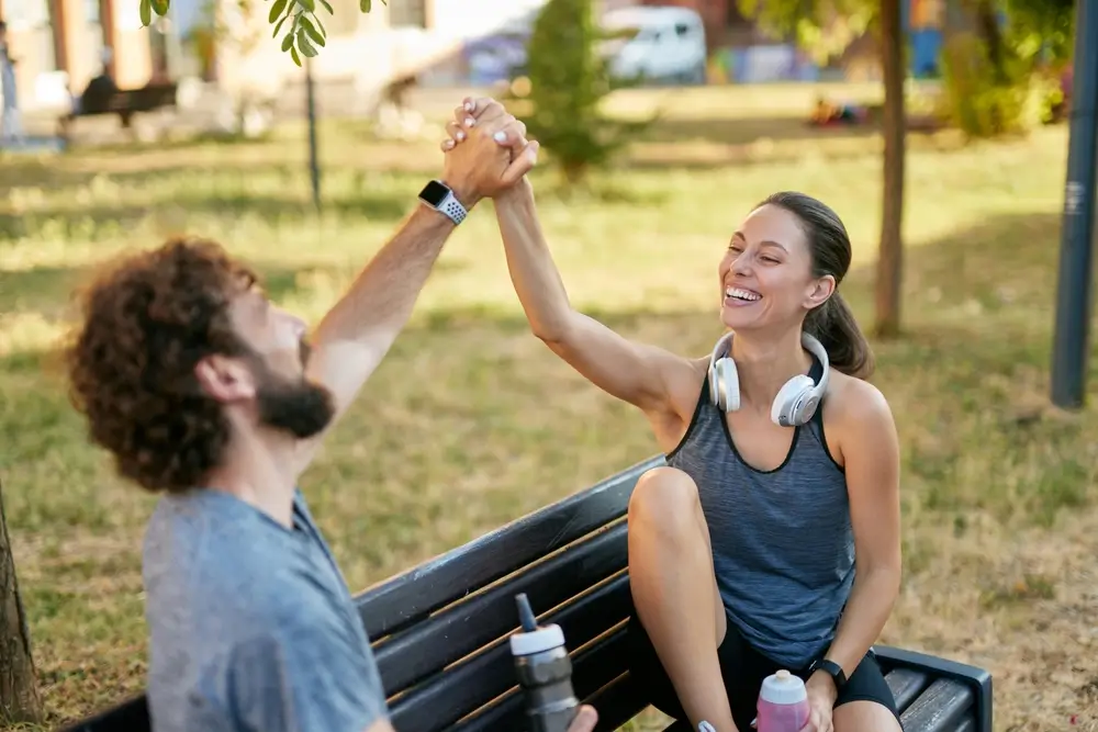 Un homme et une femme après une séance d'entraînement dans un parc, tous deux souriants assis sur un banc, mettant l'accent sur leur plaisir de remise en forme et d'amitié dans un cadre naturel.