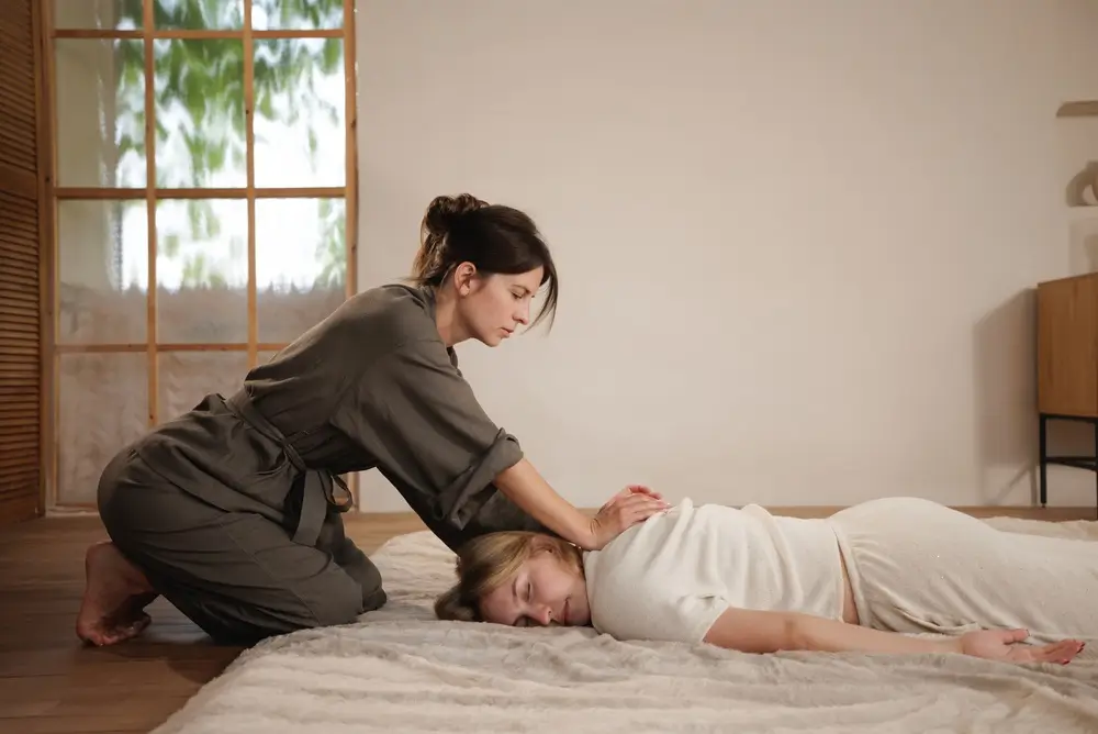 Une femme thérapeute effectue un massage traditionnel sur une cliente détendue allongée sur un tapis de sol mou dans un espace de bien-être à la maison calme.