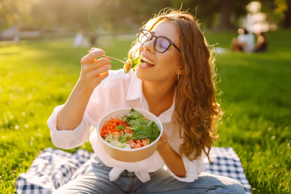 Femme mangeant un repas sain assise sur une couverture dans un parc ensoleillé.